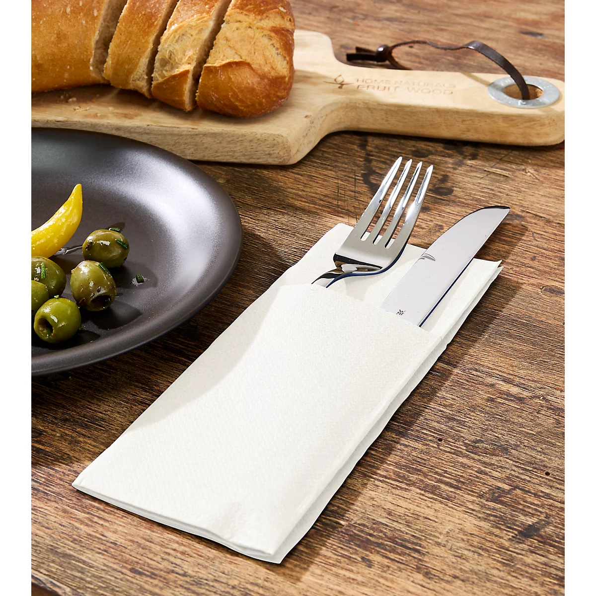 Bestek in papieren zakje op houten tafel. Bord met olijven links, brood en snijplank op de achtergrond.