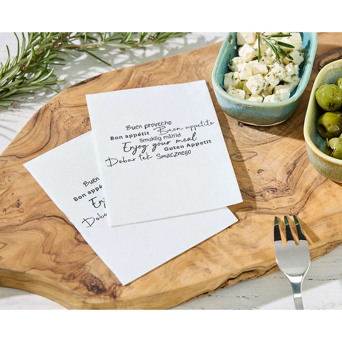 Twee servetten met de tekst 'Buen proveche, Bon appétit, Enjoy your meal' op een houten plank. Olijven, kaas en een vork.