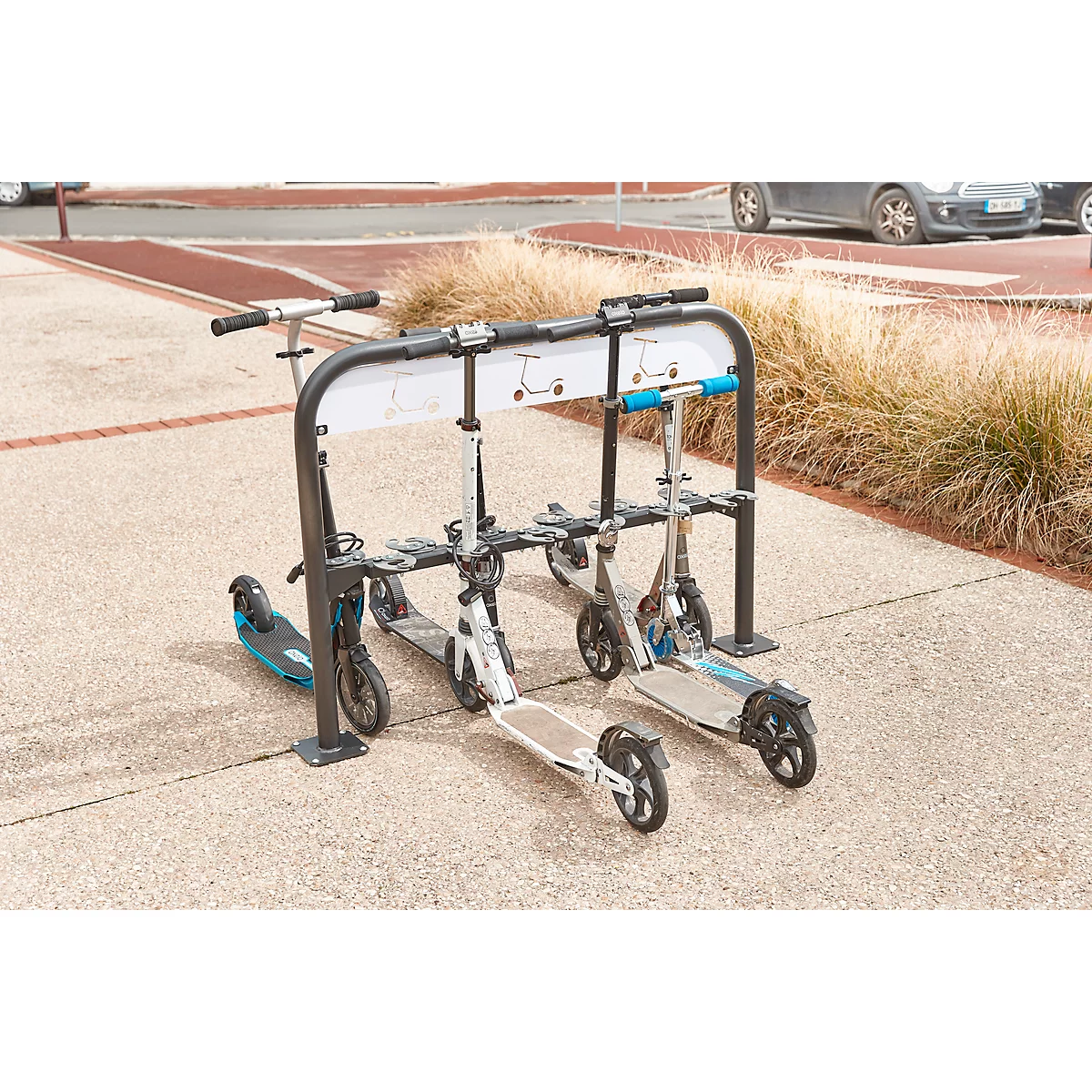 Scooters parked in a stand. Behind them is a sidewalk, to the right dry grass and in the background cars.