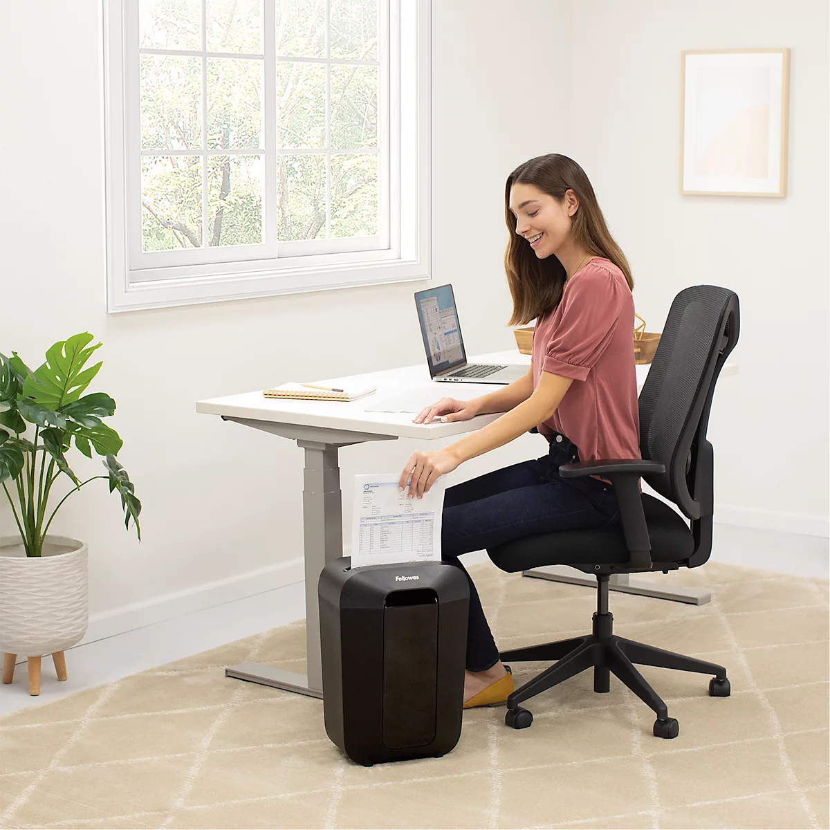 A smiling woman shreds paper at a desk. A black shredder is on the right.