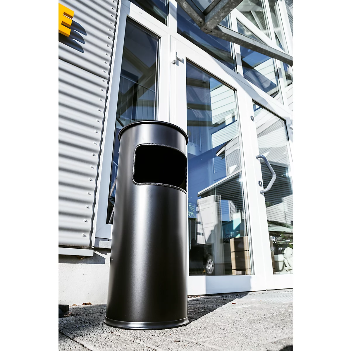 Black trash can in front of glass facade. The bin has a rectangular opening. Buildings and sky can be seen in the background.