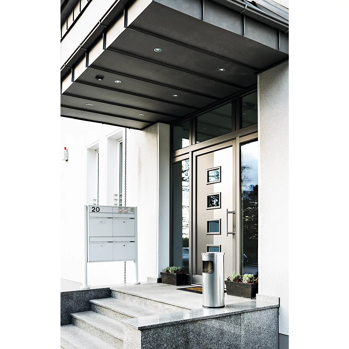 Entrance area of a building with glass front and door, steps and a small metal trash can in front of it.