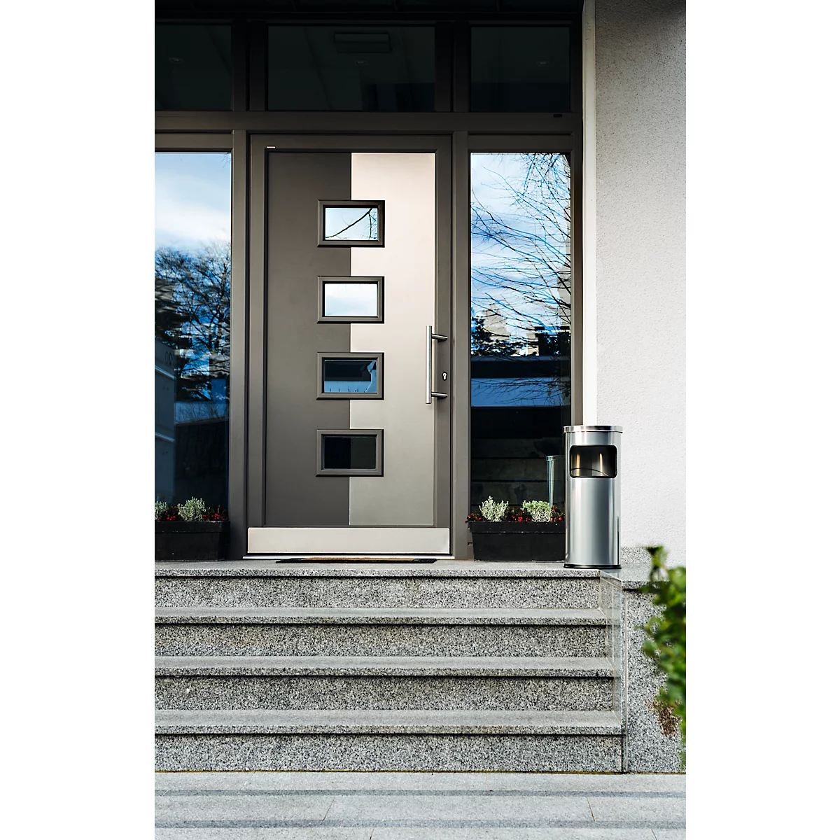 Grey front door with rectangular windows, in front of grey stone steps. Next to it, a silver trash can and flower boxes.