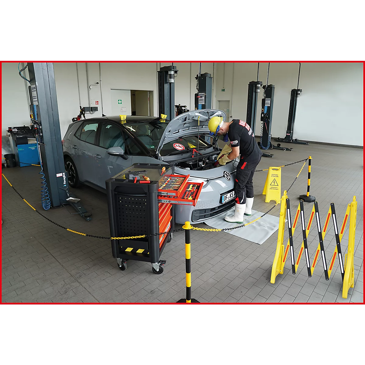 Ein Mechaniker repariert ein graues Auto in einer Werkstatt. Er trägt eine gelbe Schutzhaube und Arbeitskleidung.