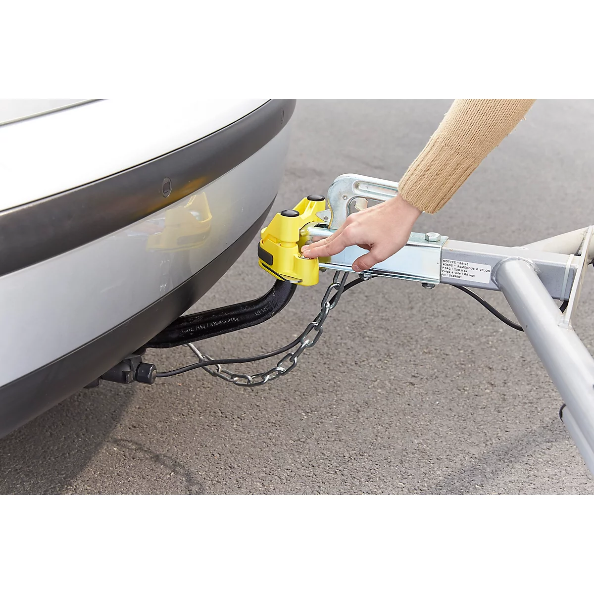 Person connecting a trailer hitch. Yellow holder attached with hand to car. Chain, cable, asphalt.
