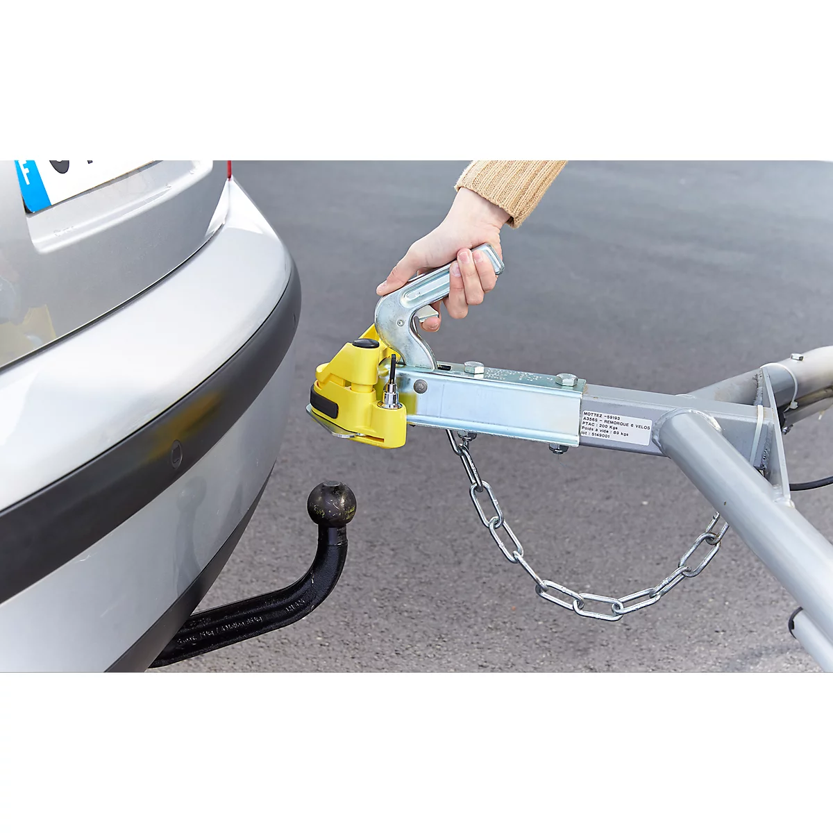 Close-up of a car with a trailer hitch. A hand is connecting the trailer hitch to a trailer.