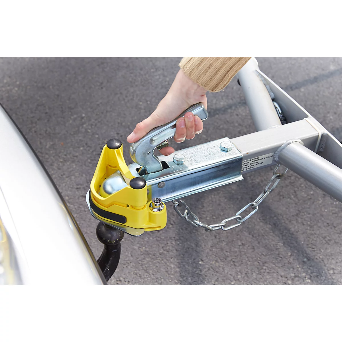 A person connecting a yellow trailer to a silver tow hitch. The hand is holding the latch.