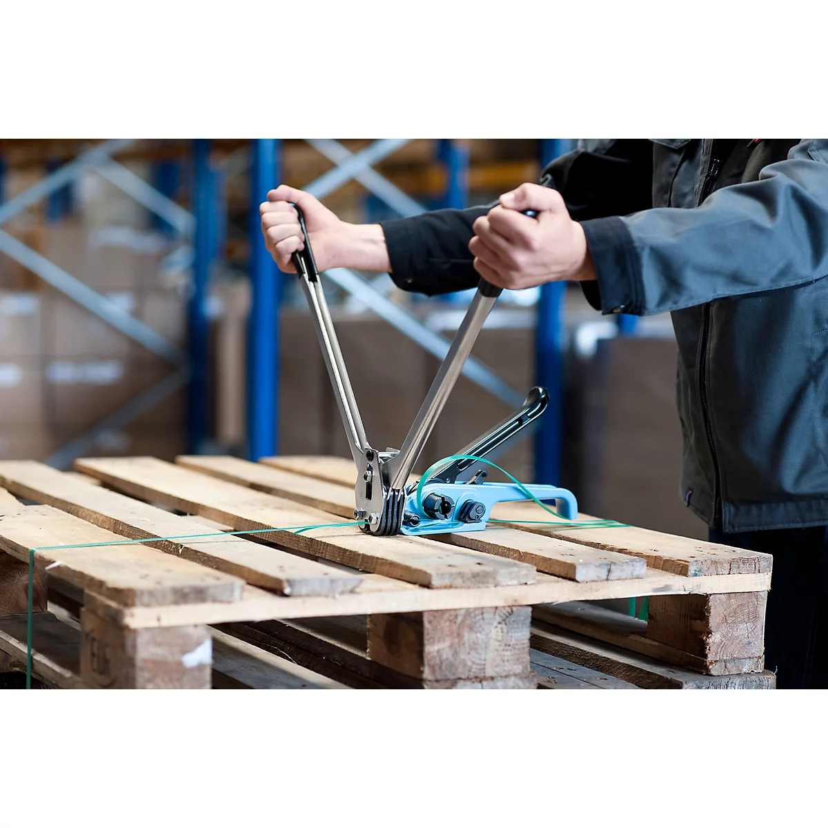 Person straps a wooden pallet with a blue tool. Warehouse background.