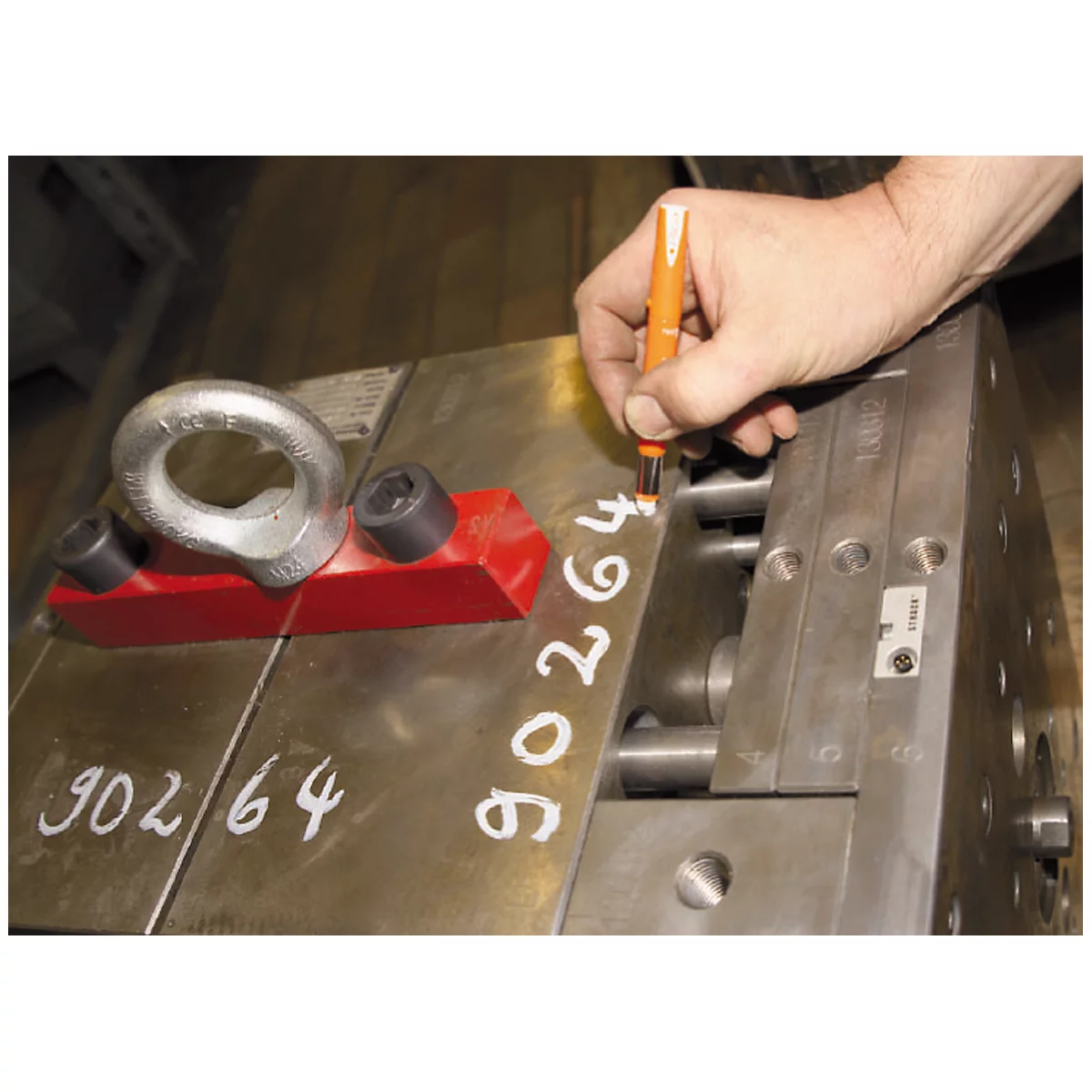 Close-up of a metal workpiece with a hand writing a number with a pen. A red block and a metal ring are attached to the workpiece.