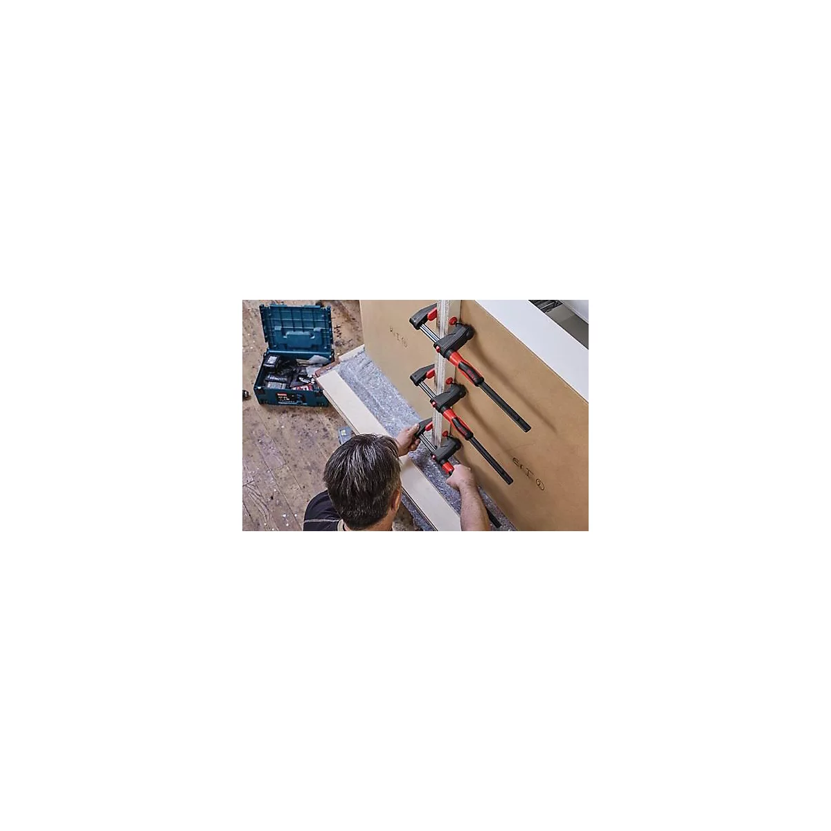 Un homme fixe deux planches de bois avec des serre-joints rouges et noirs. Une boîte à outils est à côté.