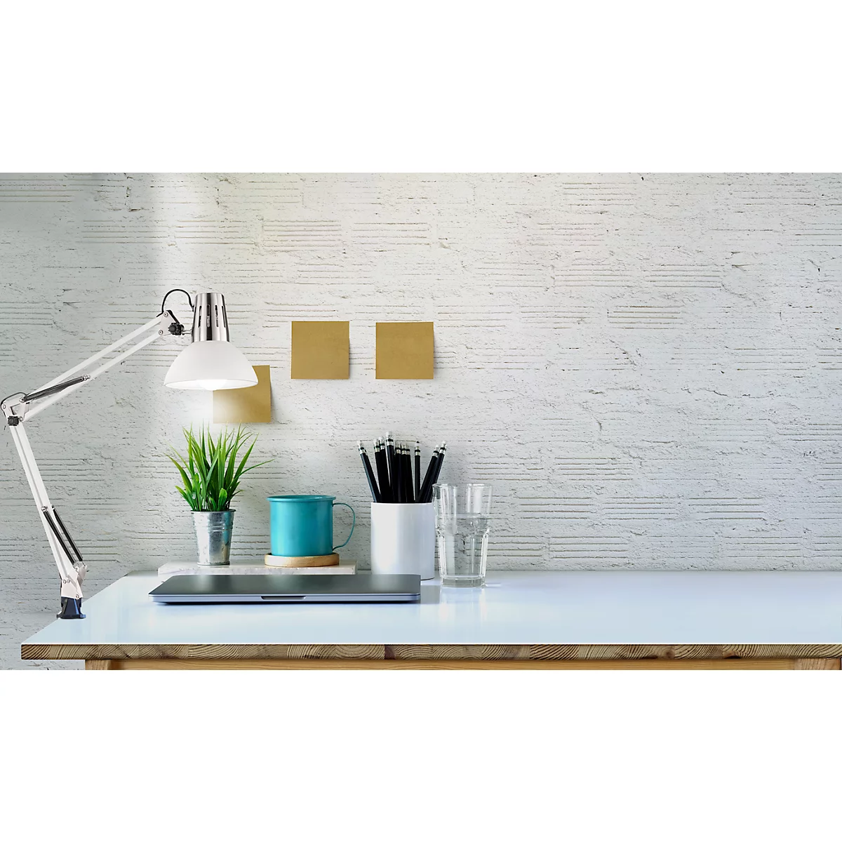 Desk with lamp, plant, pens, mug and laptop in front of a white brick wall.