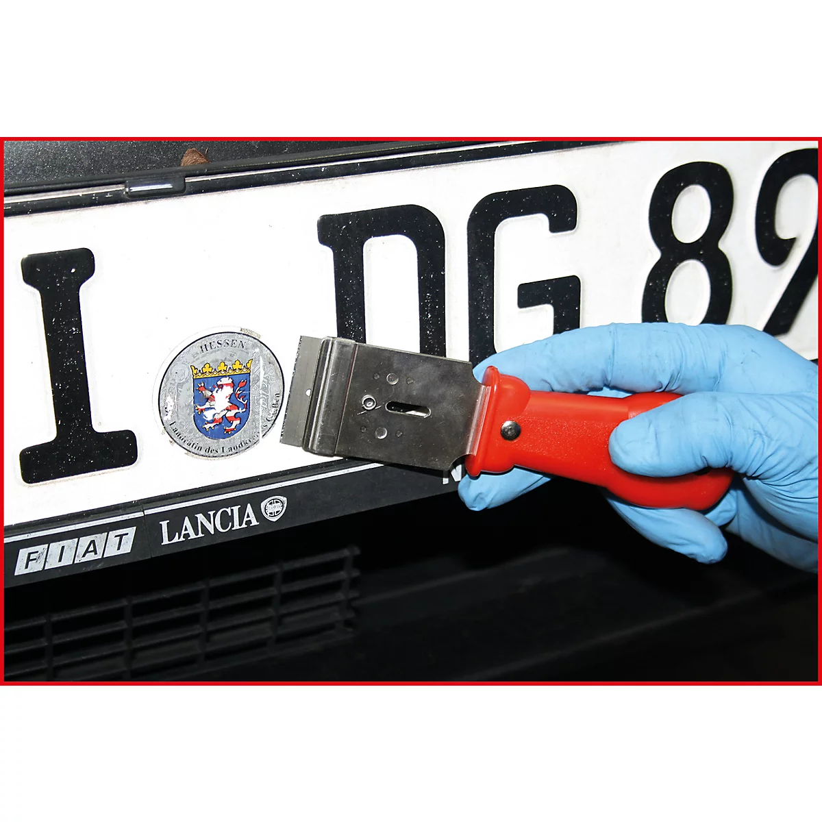 Hand mit blauem Handschuh hält Schaber an Kfz-Kennzeichen mit "Hessen"-Logo. Rotes Griffstück, Kennzeichen mit Schriftzügen, schwarzer Untergrund.