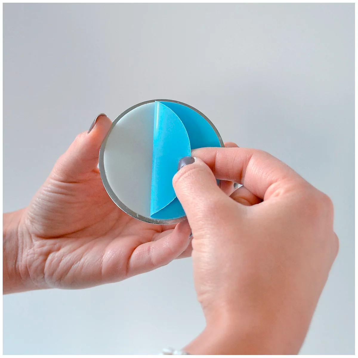 Des mains tiennent un disque rond et transparent, en train de décoller un film protecteur bleu. Le fond est blanc.