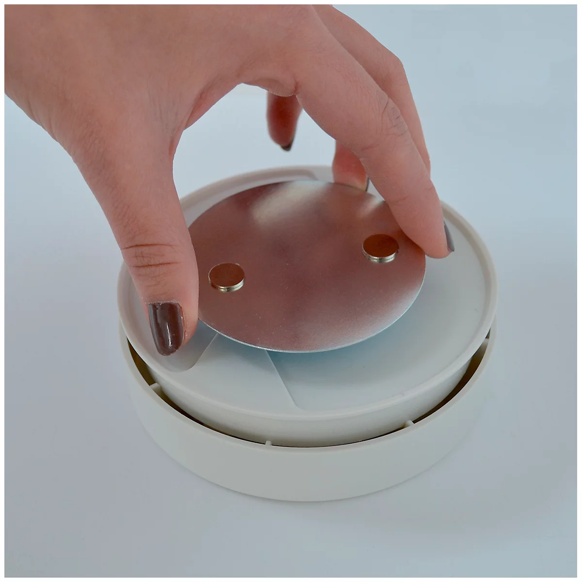 Une personne place une plaque métallique sur un objet rond blanc. Plaque avec deux aimants, main avec vernis à ongles foncé.