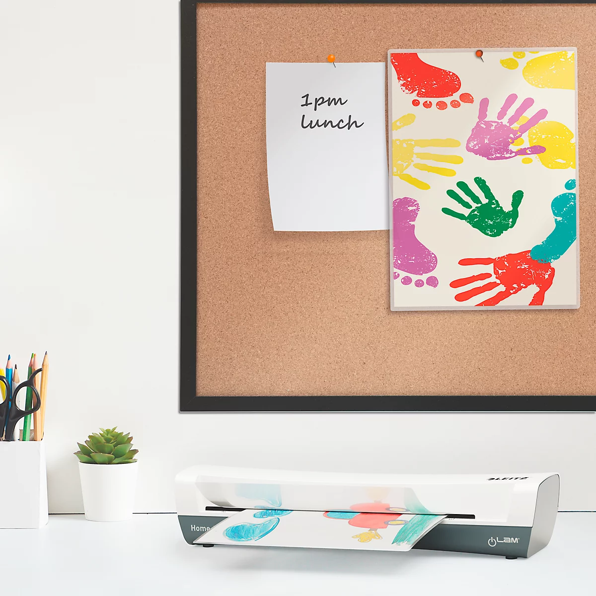 Une plastifieuse est posée sur un bureau blanc. Une note avec "13h déjeuner" et une image avec des empreintes de mains et de pieds colorées sont accrochées sur le tableau.