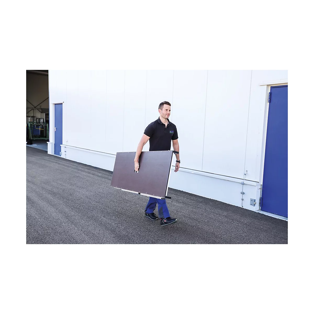 Un homme porte un plateau de table brun à l'extérieur. Il marche le long d'un mur de bâtiment blanc. Sur la gauche se trouve une porte bleue.
