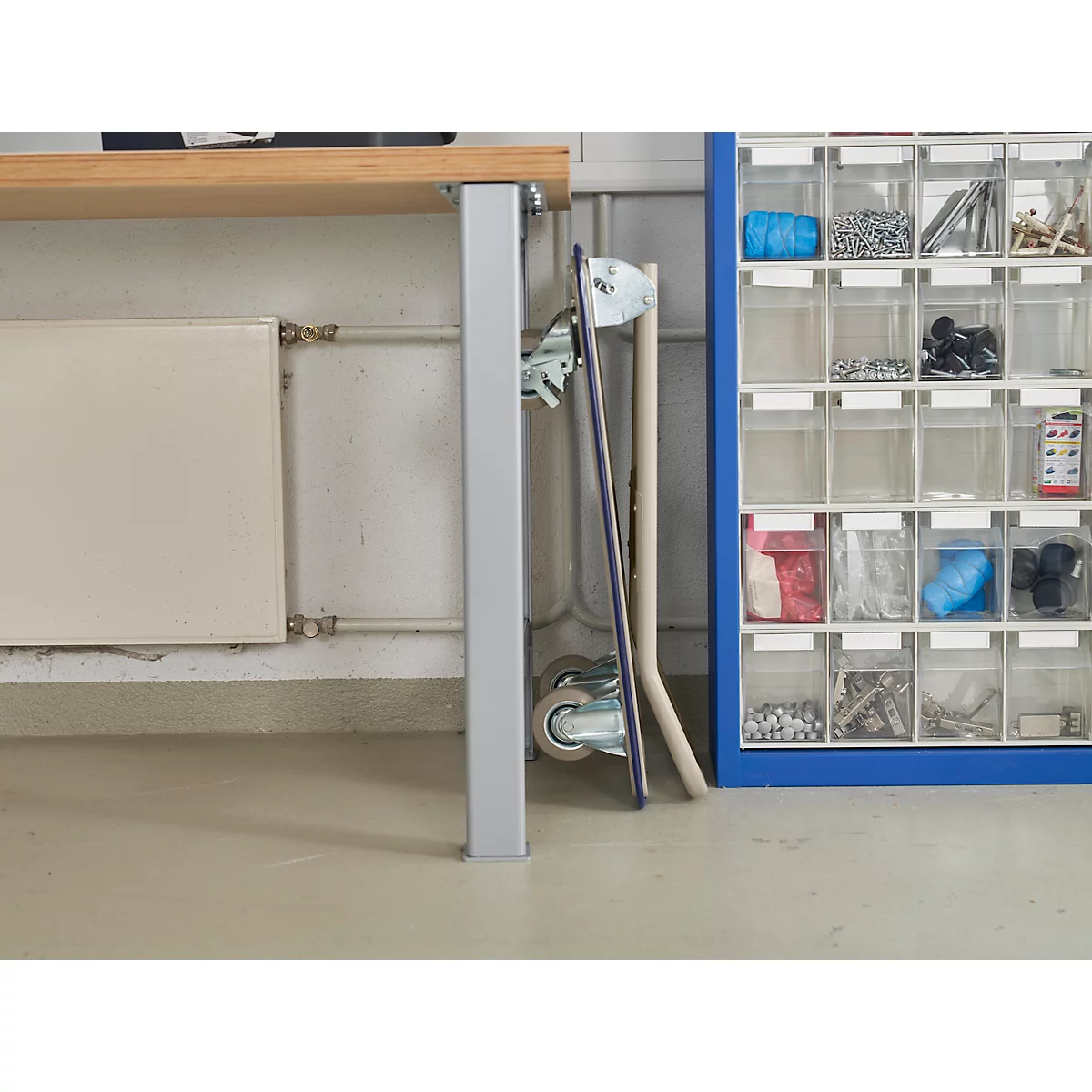 Grey workbench with wheels, next to it a blue shelf wall with small parts in transparent boxes.