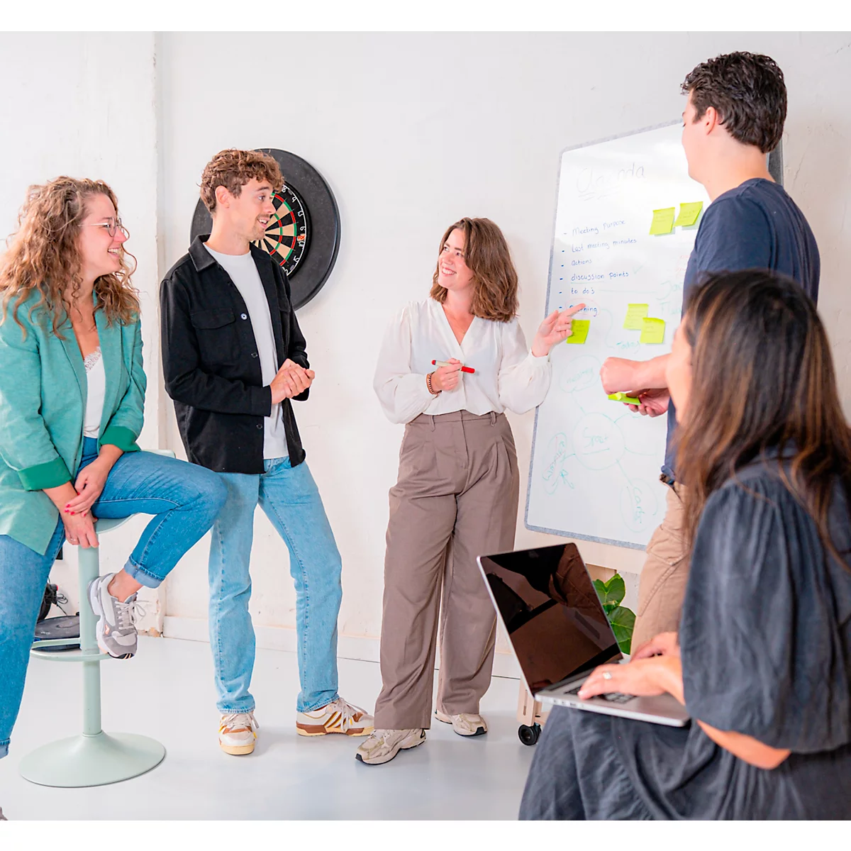 Un groupe de jeunes discutent devant un tableau blanc avec des notes. Certains tiennent des stylos et un ordinateur portable est au premier plan.