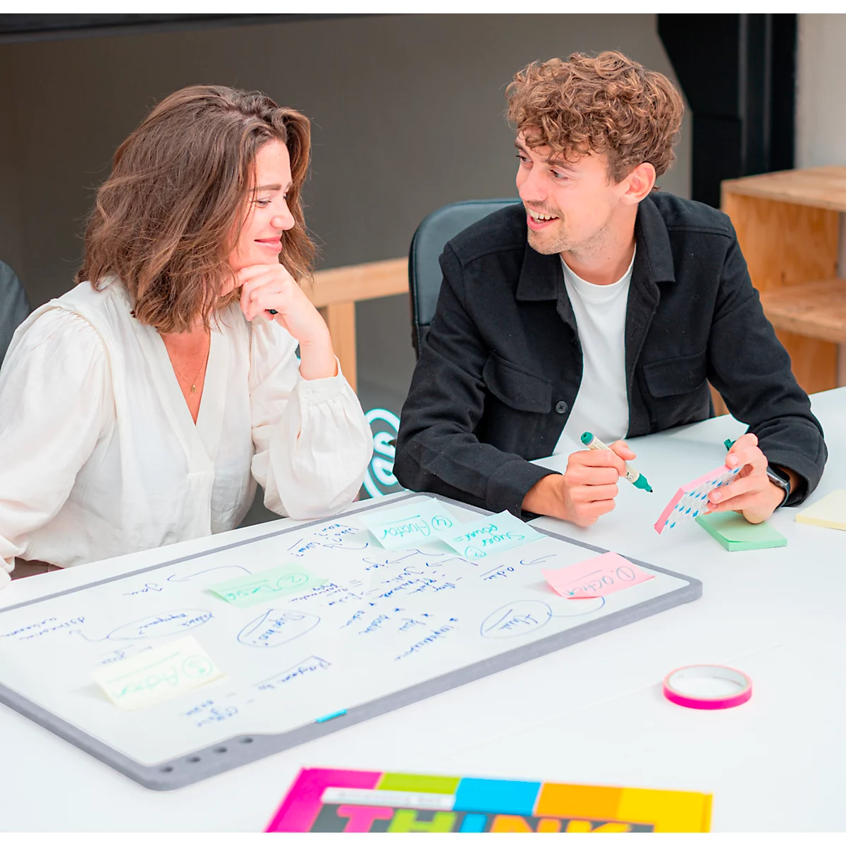 Deux personnes regardent un tableau blanc avec des notes et des dessins. Ils ont l'air concentrés et interagissent.