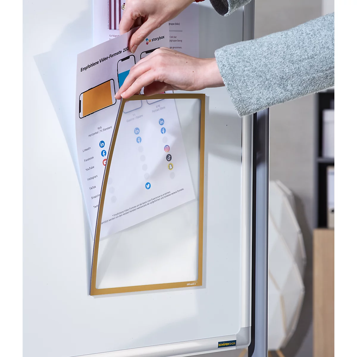 Des mains placent du papier dans un cadre doré. Un tableau blanc est visible en arrière-plan.