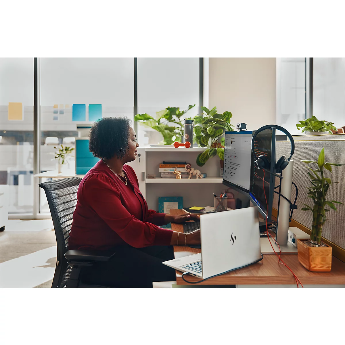 A woman sits at a desk in front of a computer. She is wearing a red top and has dark hair. There is a laptop and potted plants on the desk.