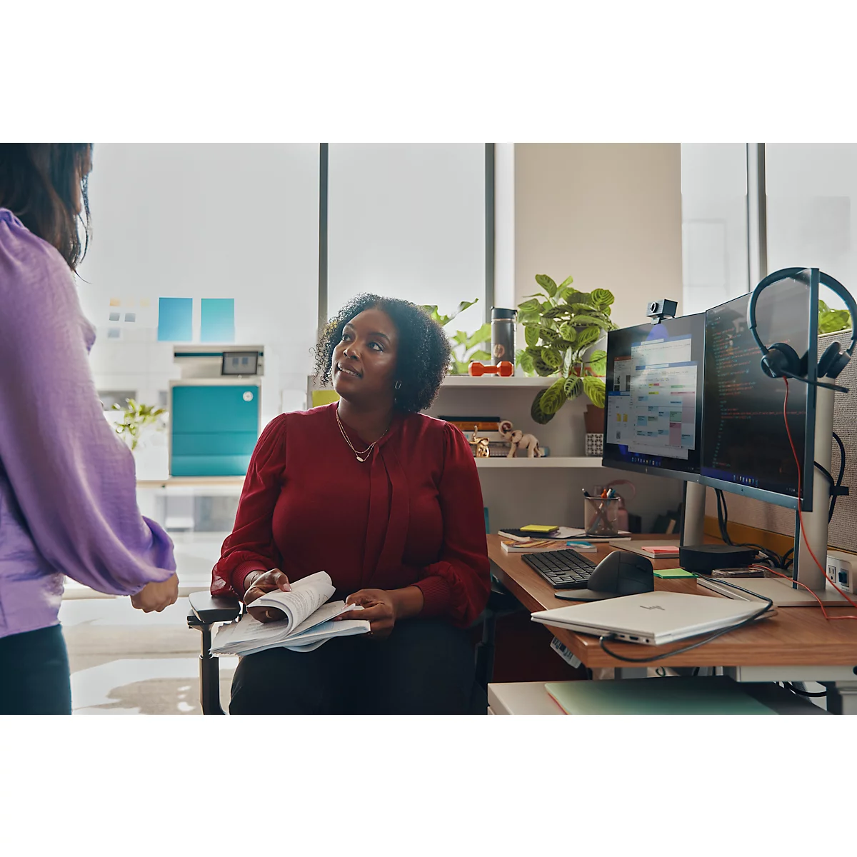 A woman in a red blouse is seated, looking up at a person standing in front of her, holding a book. A desk with computers is visible.