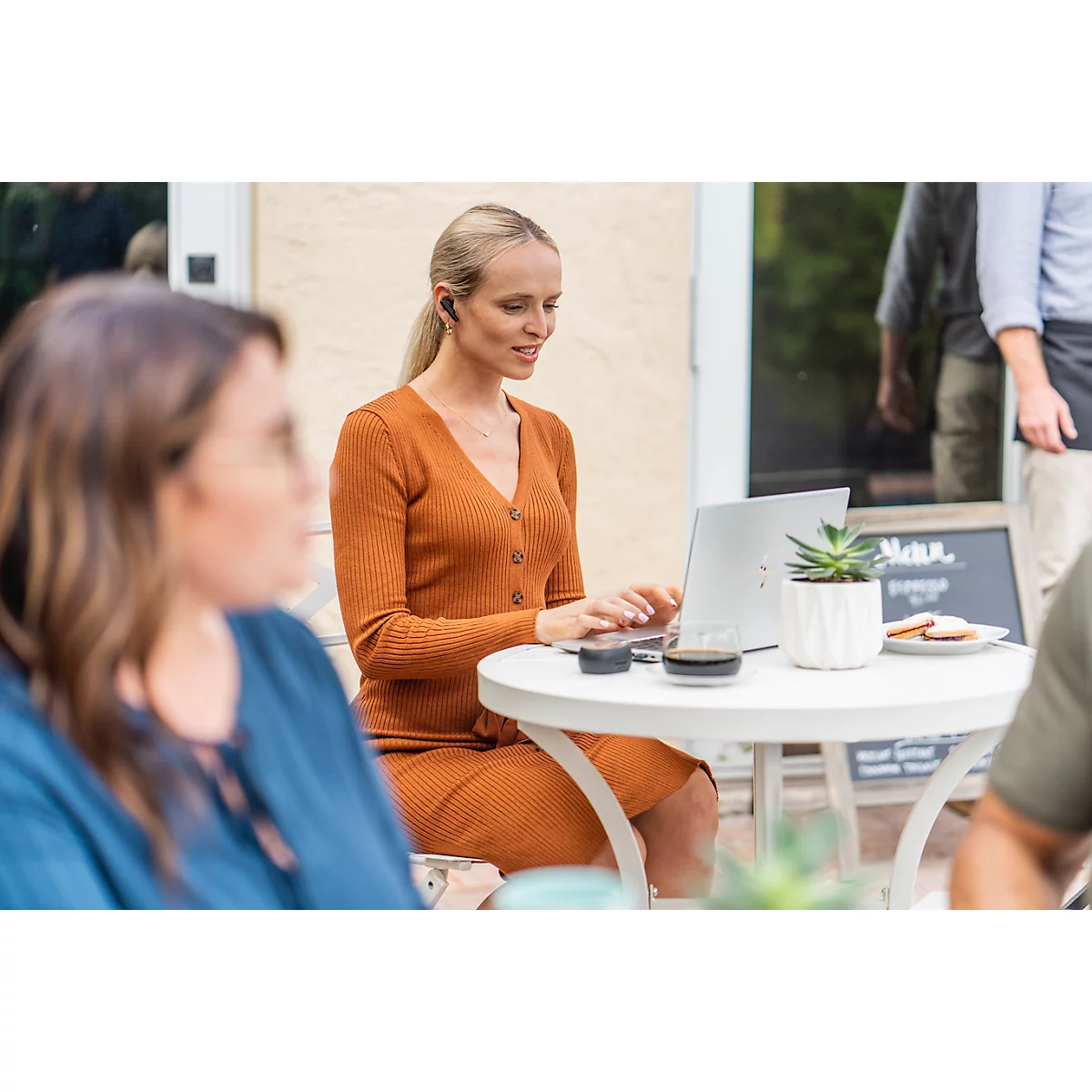 A blonde woman working on a laptop at a small table outdoors, wearing earbuds and a brown dress.