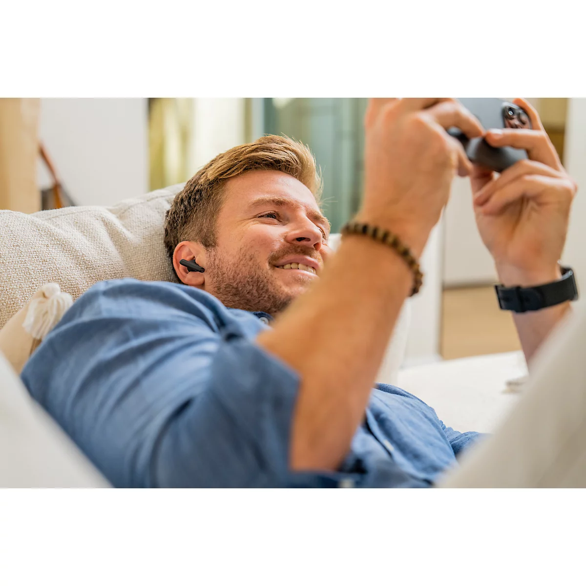 A man with blond hair lies relaxed on a sofa, looks at his smartphone and wears earbuds.