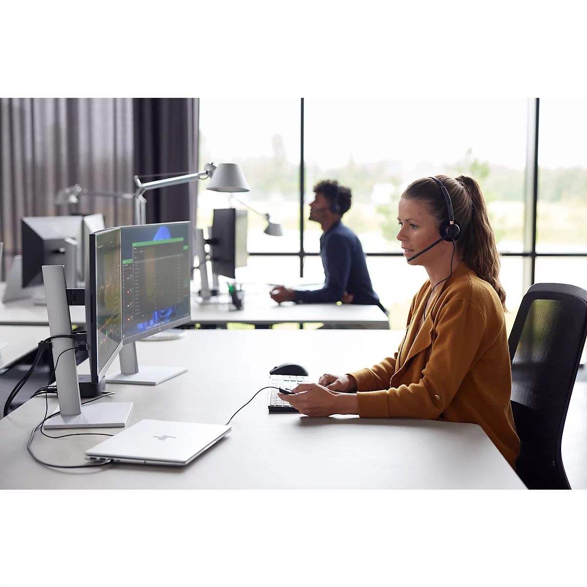 A woman wearing a headset works at a computer in an office. A man in the background, also wearing a headset.