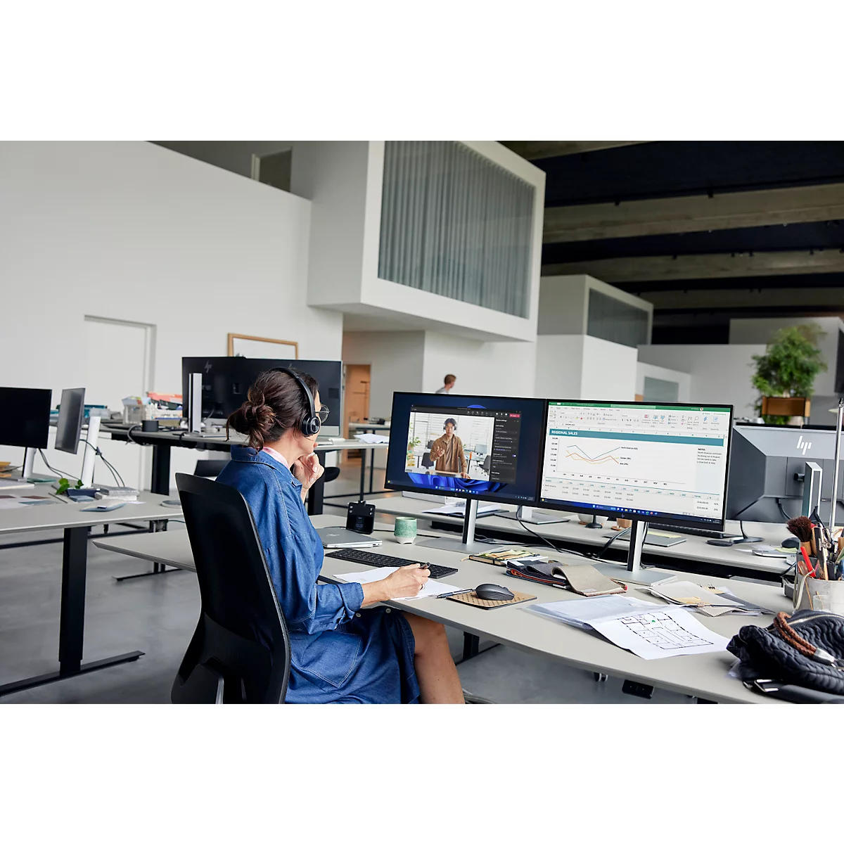 A woman sits at a desk in an office. She is wearing a headset and looking at two monitors that show video conferencing and spreadsheets.