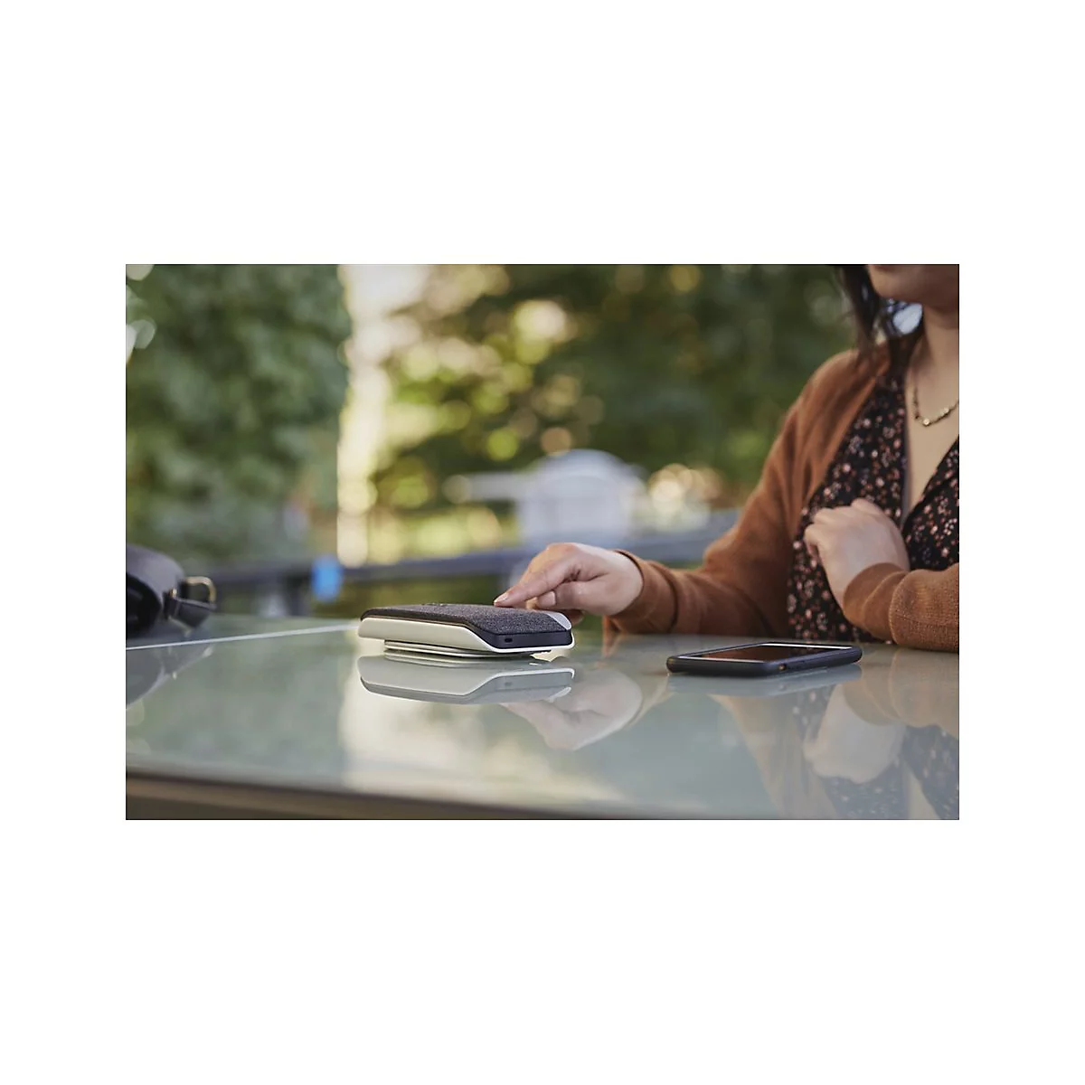 Eine Frau legt ihr Smartphone neben einen Lautsprecher auf einen Tisch. Im Hintergrund ist ein Baum unscharf.