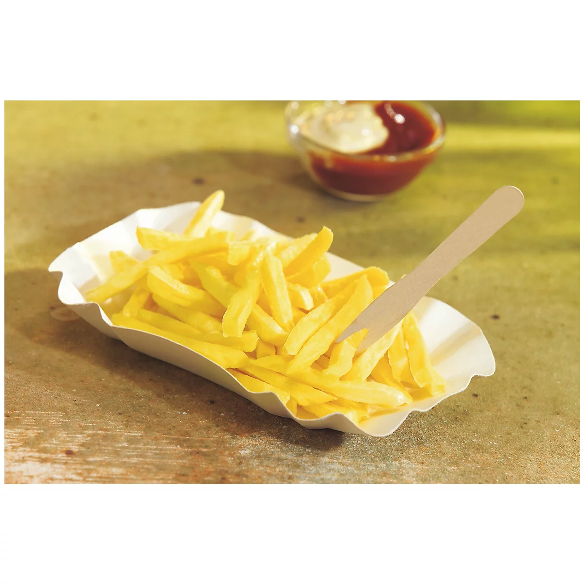 Pommes Frites in einer Papierschale mit Ketchup und Mayo im Hintergrund. Ein Holzbesteck steckt im Essen.
