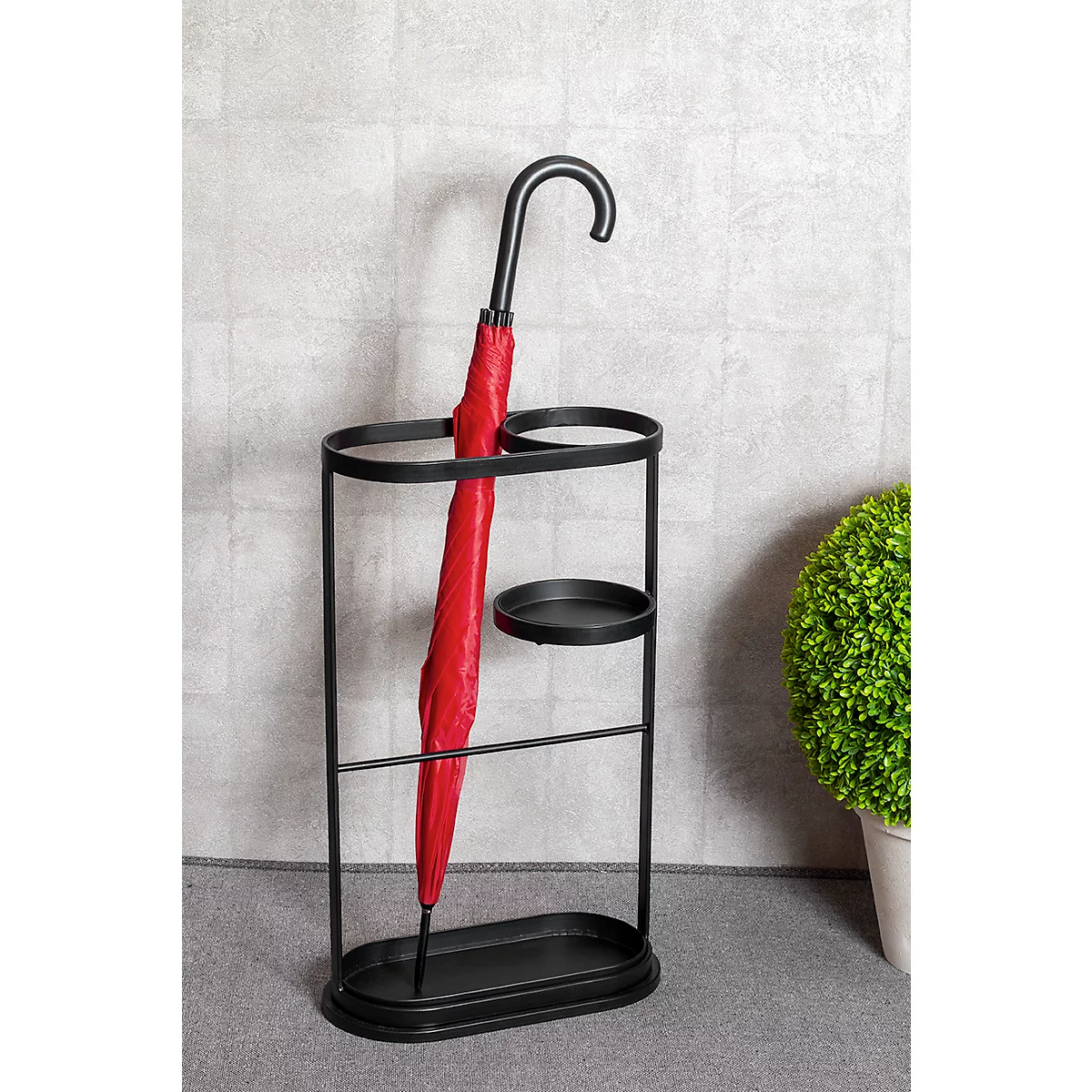Porte-parapluie noir avec parapluie rouge, fond gris, plante verte à droite.