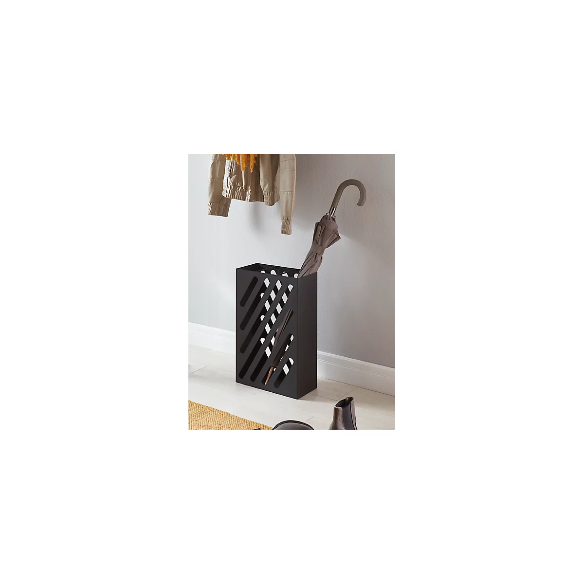 Porte-parapluie noir avec fentes diagonales. Un parapluie est à l'intérieur. Une veste est suspendue au mur.