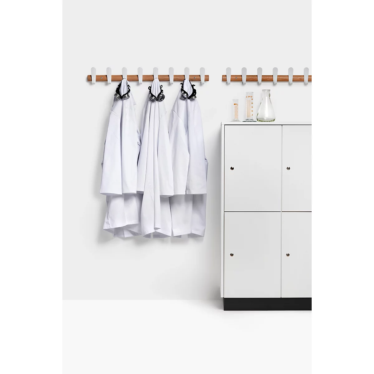 Des blouses blanches de laboratoire sont accrochées à un porte-manteau en bois et métal devant une armoire blanche.