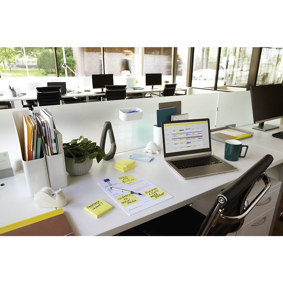 Espace de travail de bureau avec ordinateur portable, notes et plantes. En arrière-plan, d'autres espaces de travail et une personne.