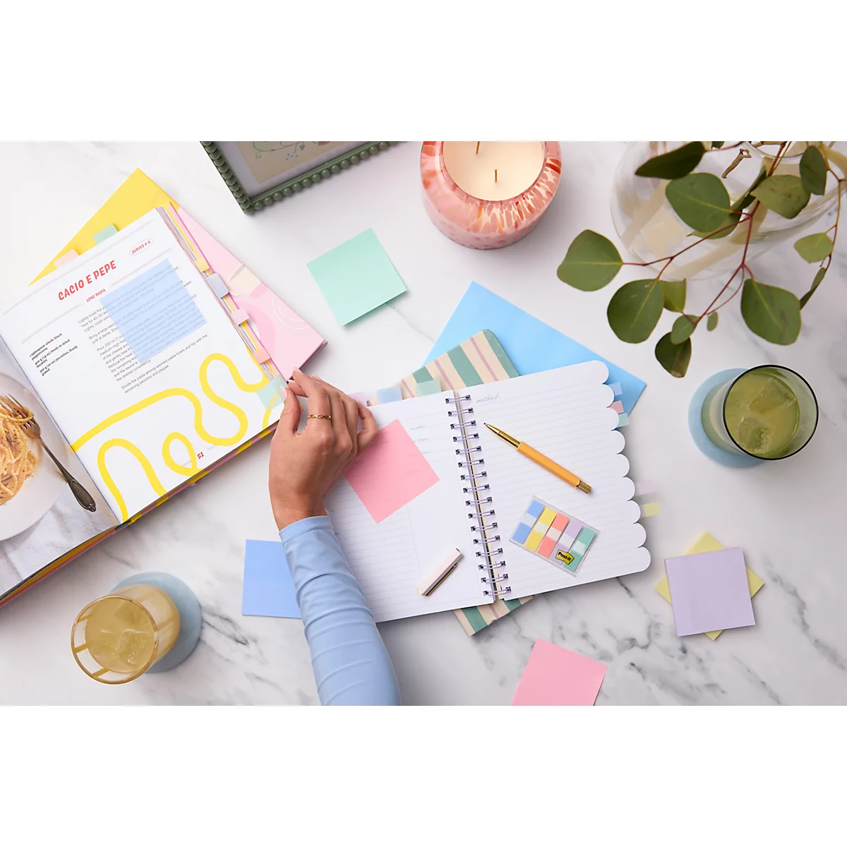 Vue aérienne d'un bureau avec un carnet, un stylo, des post-it, un livre et des boissons. Une main écrit.