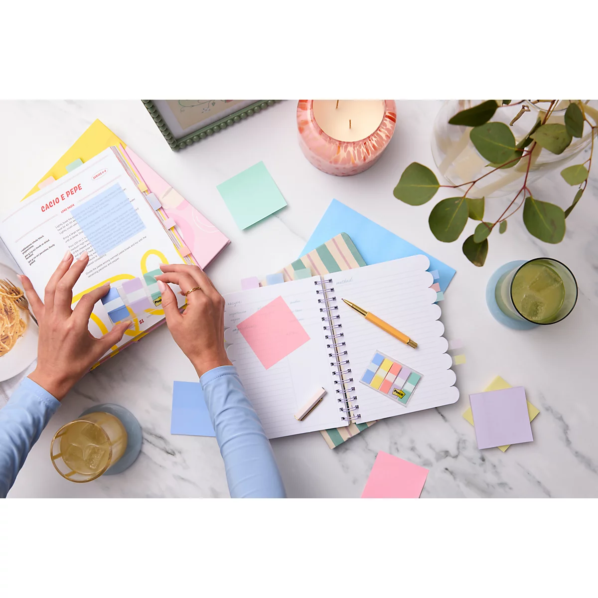 Vue de dessus d'une main prenant des notes dans un cahier. Des stylos, des post-it et des boissons sont éparpillés autour.