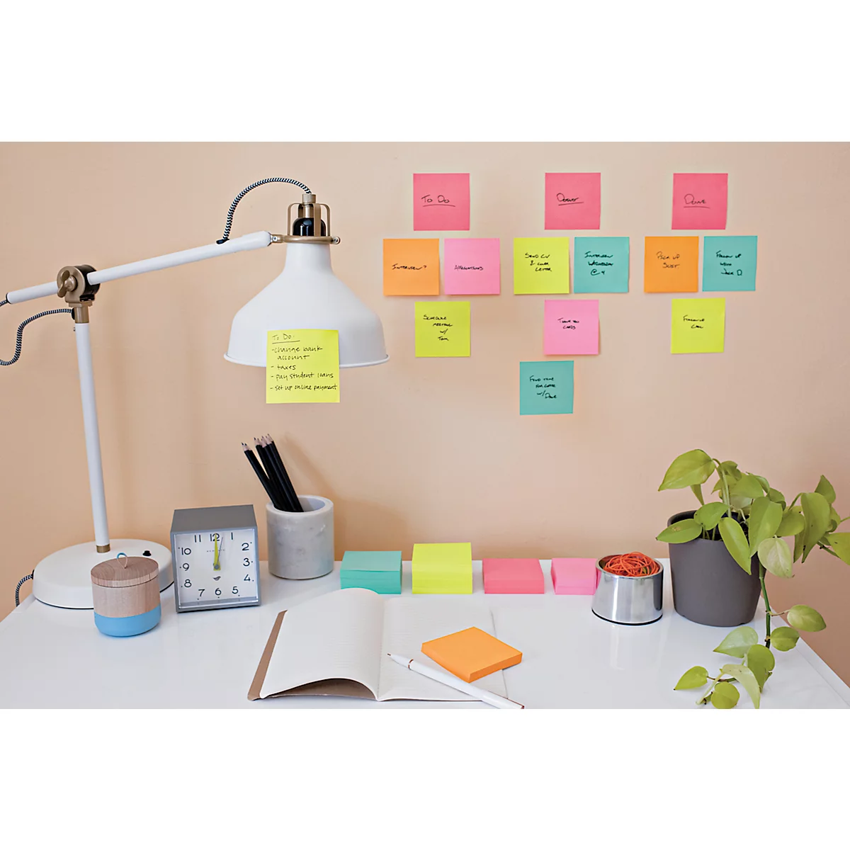 Schreibtisch mit Lampe, Uhr, Notizzetteln und Pflanze. Einige Notizzettel mit handschriftlichen Notizen an der Wand.