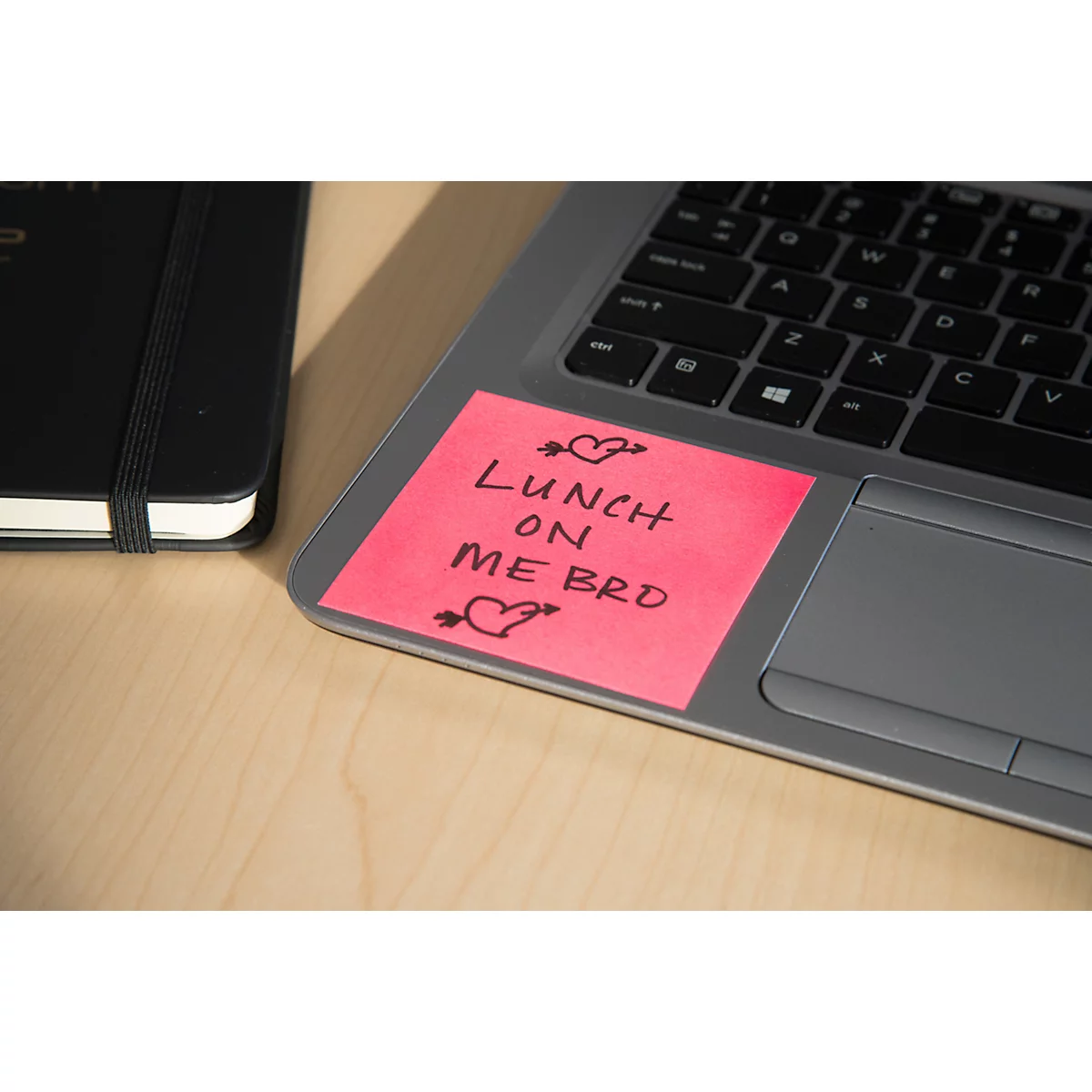 Ein pinkfarbener Notizzettel mit der Aufschrift „Lunch on me bro“ klebt auf einem Laptop, daneben ein schwarzes Notizbuch.