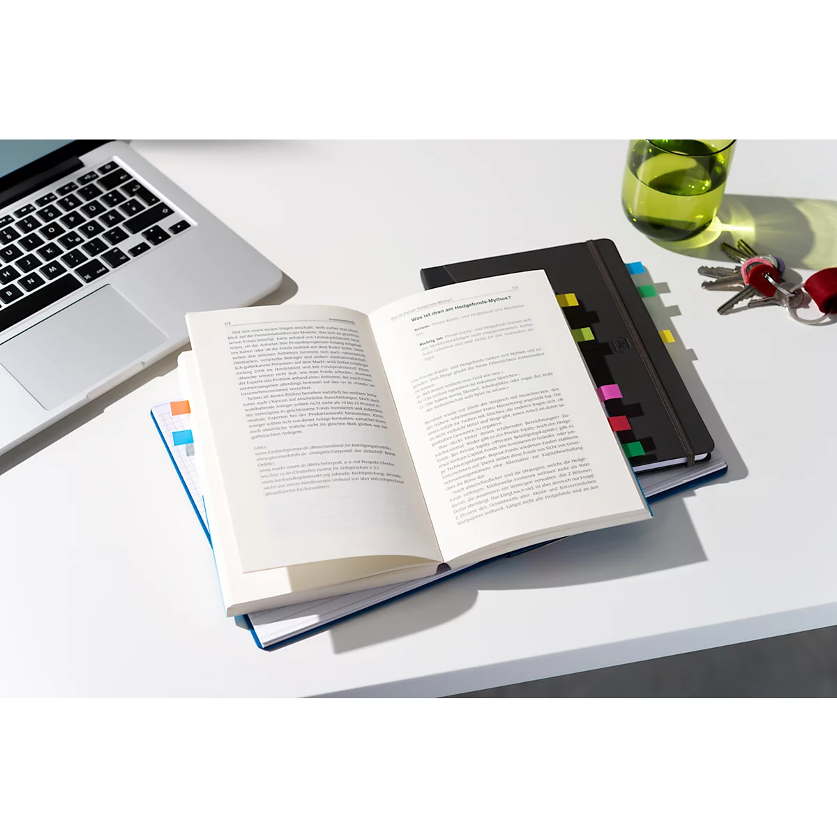 Livre ouvert avec des notes adhésives colorées, un ordinateur portable, un verre et des clés sur une table blanche.