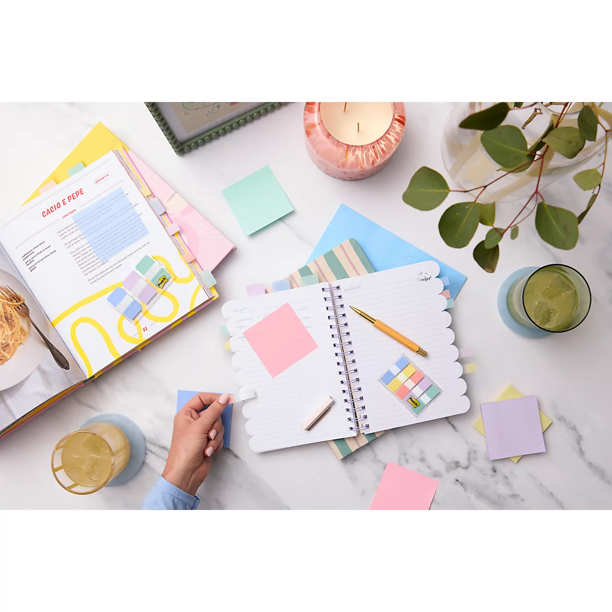 Bovenaanzicht van een bureau met notitieboek, pennen, plaknotities, drankjes en kookboek. Een hand plaatst een plaknotitie.