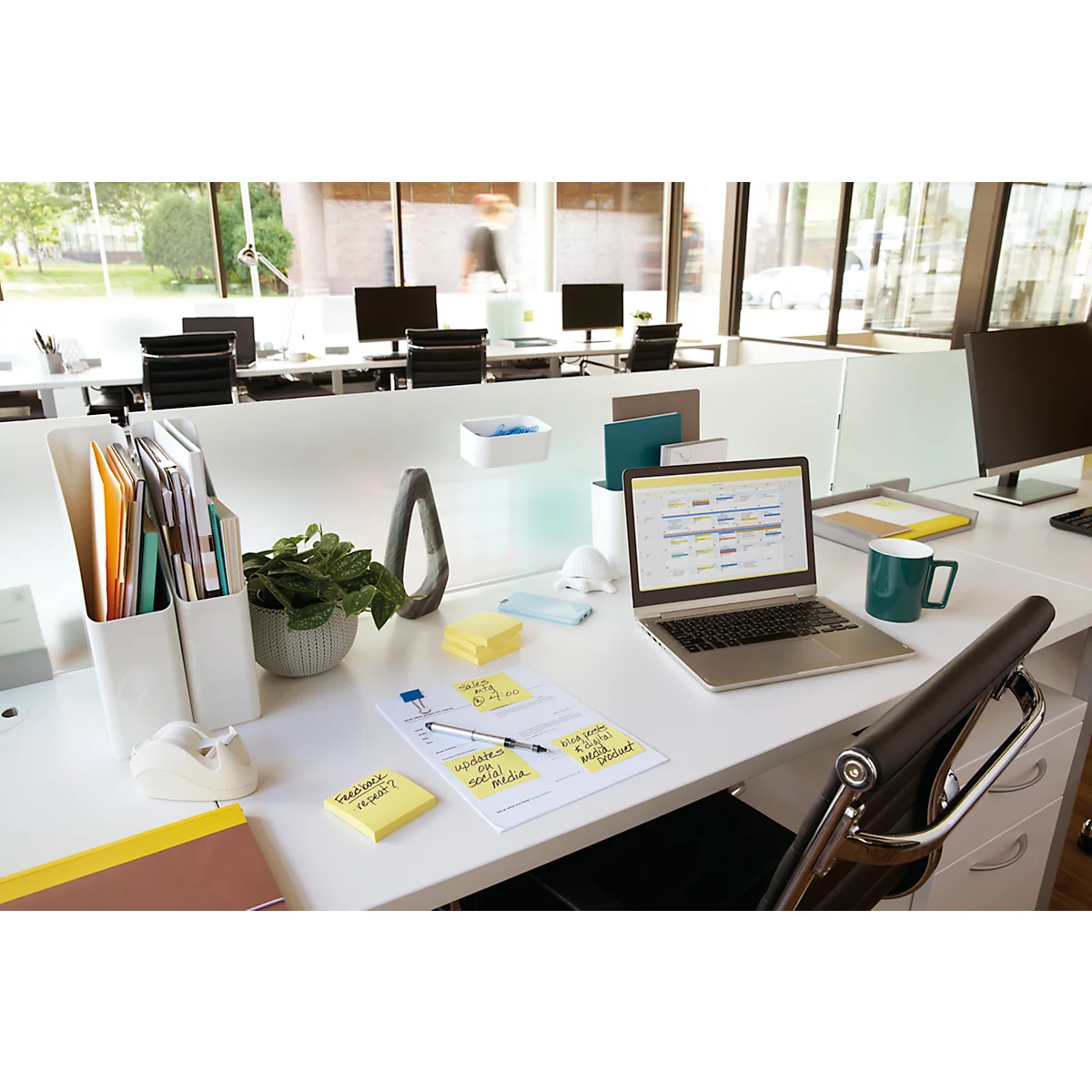 Bureau met laptop, notities, pennen, groene mok en plant. Andere werkplekken op de achtergrond.