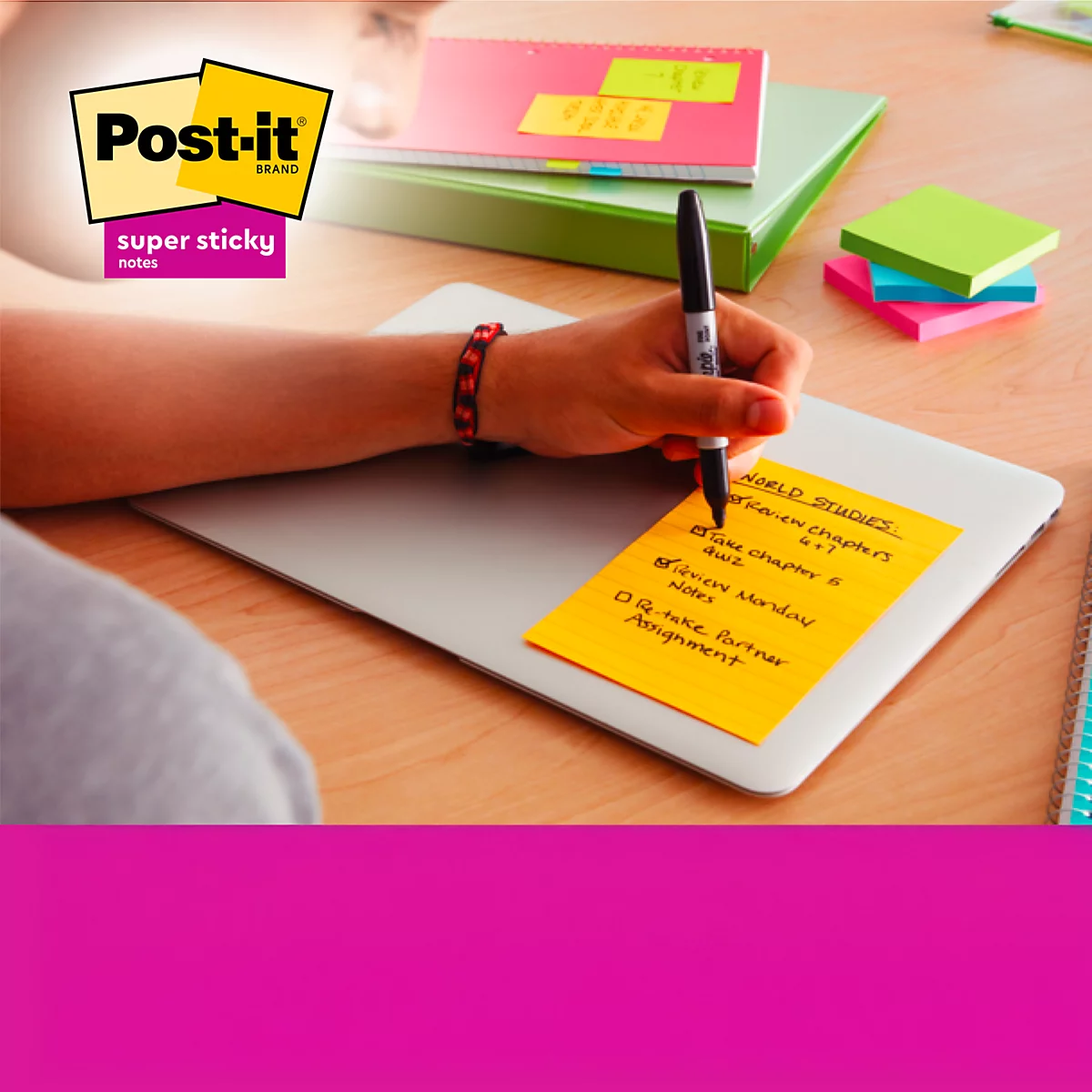 Persoon schrijft met zwarte pen op gele Post-it, vastgeplakt op een laptop. Meer notities en blokken op de achtergrond.