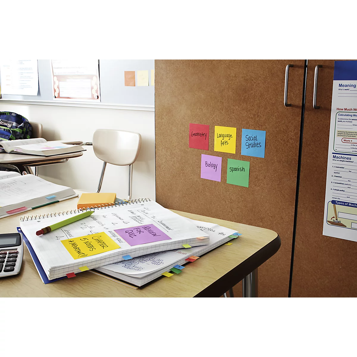 Desk with notebook, pen, sticky notes and cupboard with colourful notes with school subjects written on them.