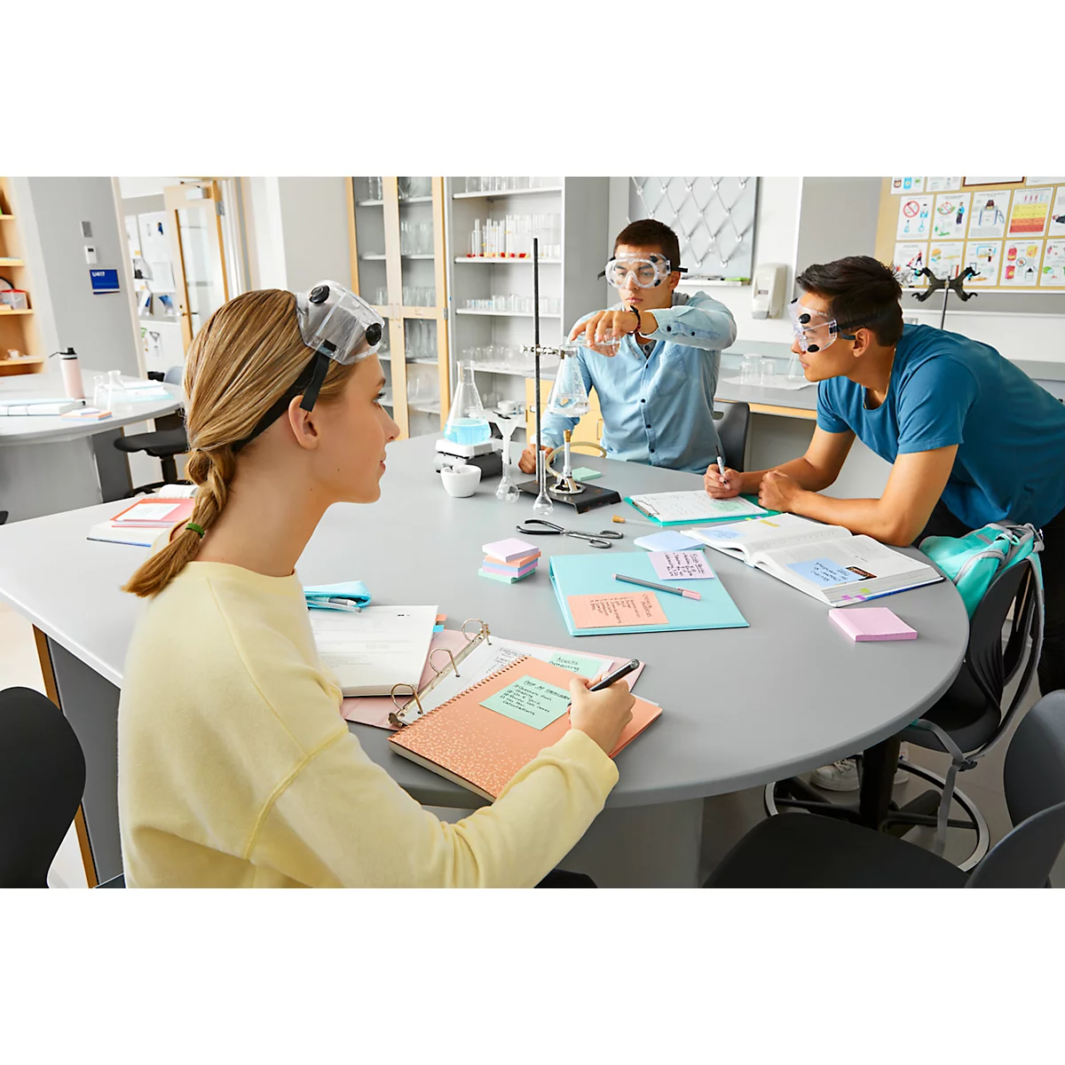 Drei Schüler in einem Labor, das Experimente durchführt. Eine Schülerin schreibt, zwei schauen zu.