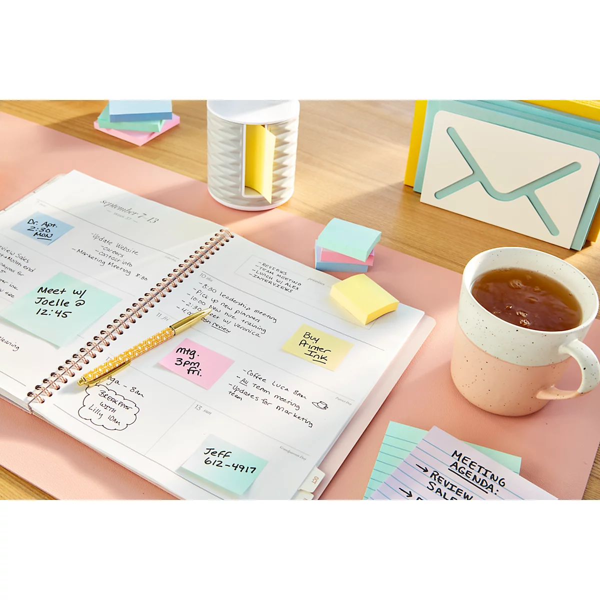 Un agenda avec des post-it, un stylo et une tasse de thé sur un bureau rose.