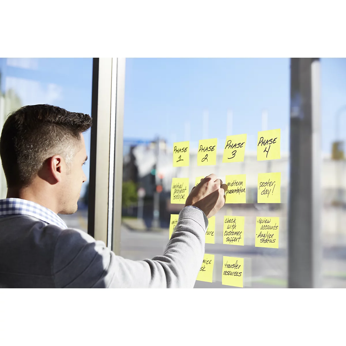 Un homme écrit sur des post-it jaunes sur une vitre, des notes sur les phases d'un projet. Vue de la ville en arrière-plan.