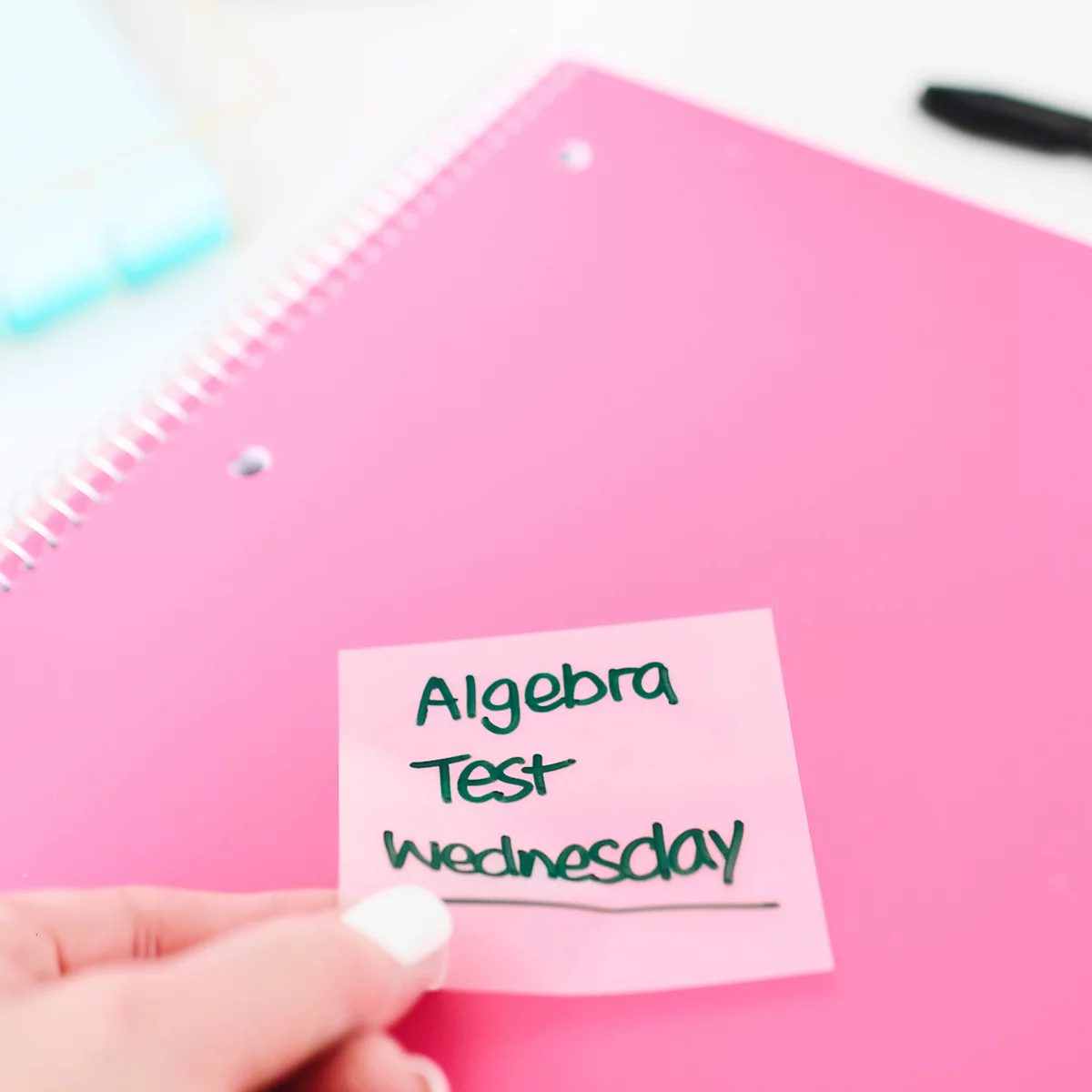 Close-up van een hand die een roze plakbriefje vasthoudt met 'Algebra Test Wednesday' op een roze spiraalgebonden notitieboek.