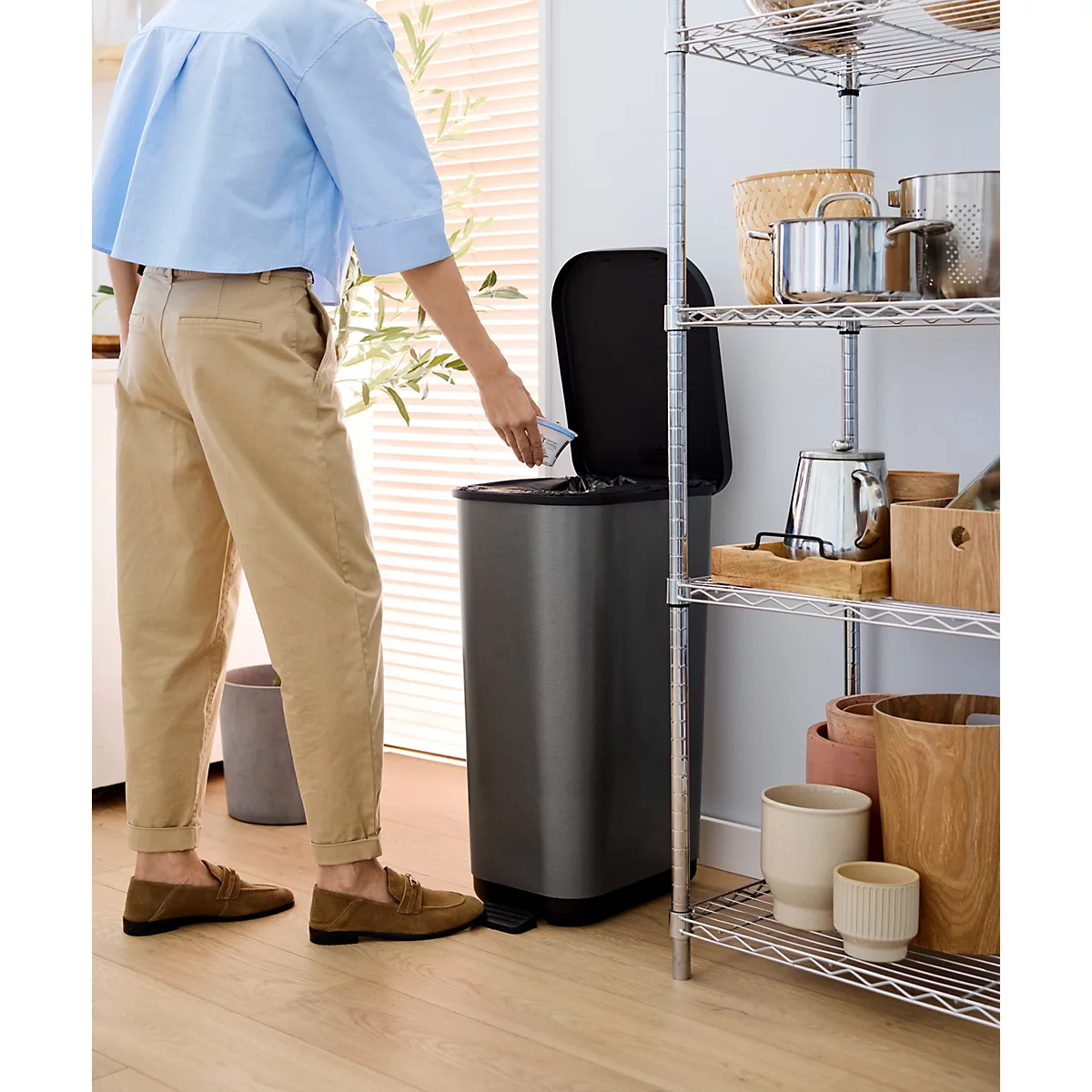Une personne jette des déchets dans une poubelle anthracite. À côté du bac se trouve une étagère avec des ustensiles de cuisine.